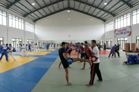 Kesibukan di aula latihan Puslatda Bela Diri Jatim, di mana atlet dari berbagai cabor seperti judo/kurash (kiri) dan kickboxing (kanan) berlatih bersama. KONI Jatim membuka opsi perekrutan lintas cabor untuk mengatasi krisis atlet di beberapa nomor pertandingan demi memenuhi target medali emas di PON Bela Diri II 2026. (foto ilustrasi AI)